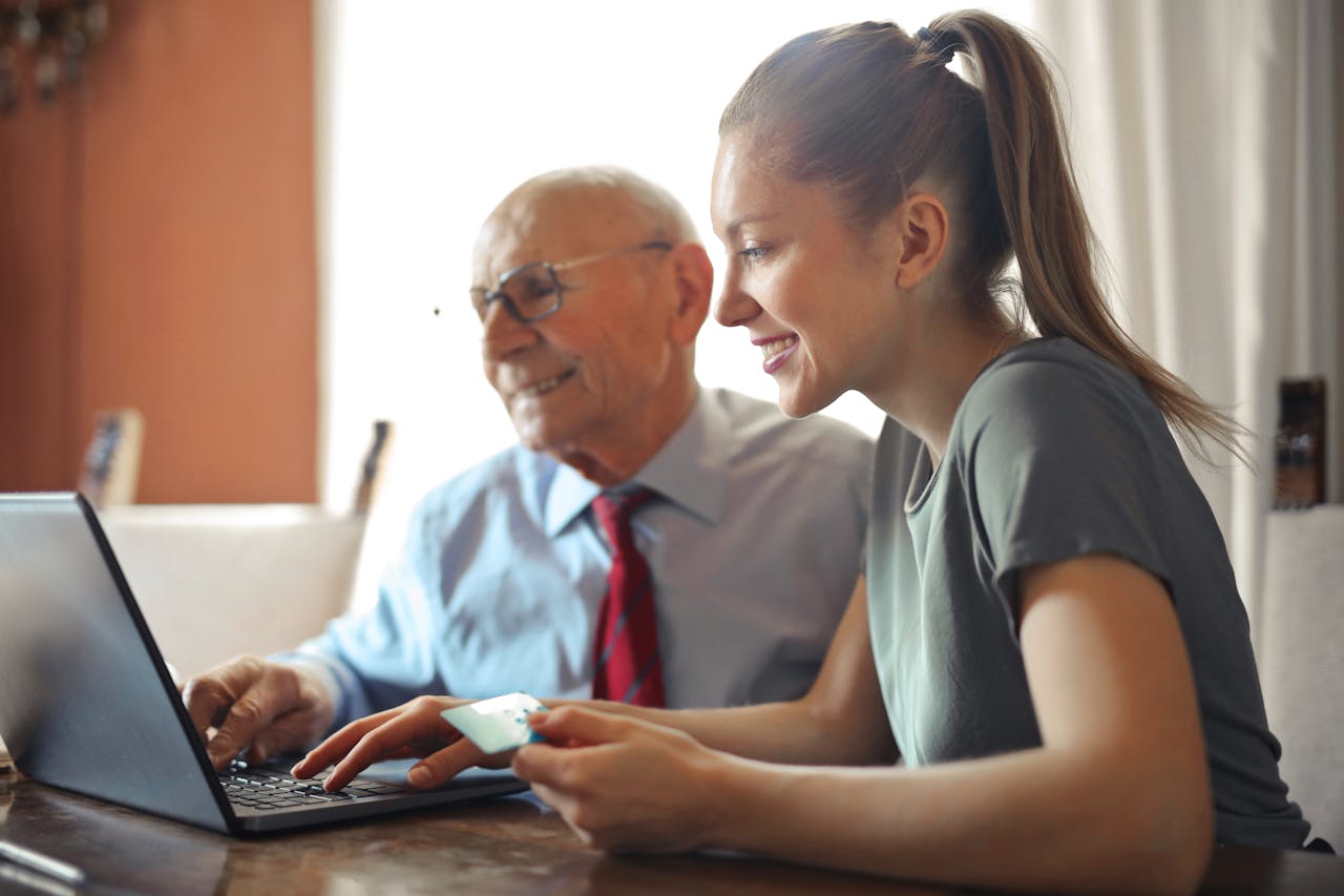 creative Young woman in casual clothes helping senior man in formal shirt with paying credit card in Internet using laptop while sitting at table